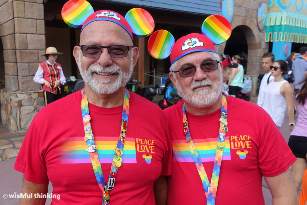 At Magic Kingdom, two older men in red t-shirts saying Peace and Love, plus rainbow Mickey Mouse ears, smile at Walt Disney World, Orlando, Florida