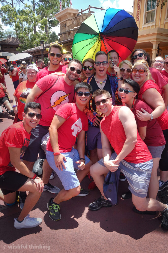 A smiling group of men, women, and non-binary LGBTQ+ friends hug and laugh with a rainbow umbrella and red t-shirts during Disney's Gay Days weekend in Orlando, Florida's Magic Kingdom
